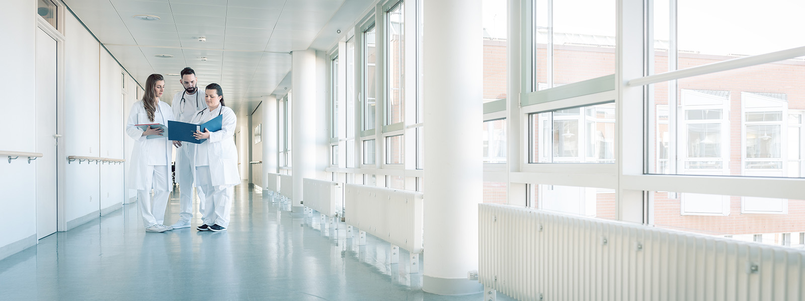 2 Ärztinnen und 1 Arzt über eine Akte gebeugt auf einem langen Krankenhausflur