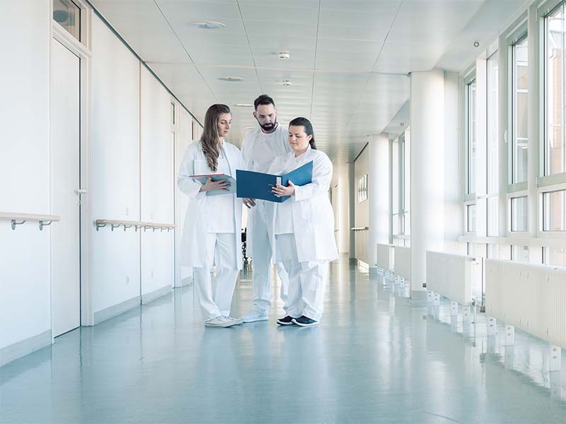 2 Ärztinnen und 1 Arzt über eine Akte gebeugt auf einem langen Krankenhausflur